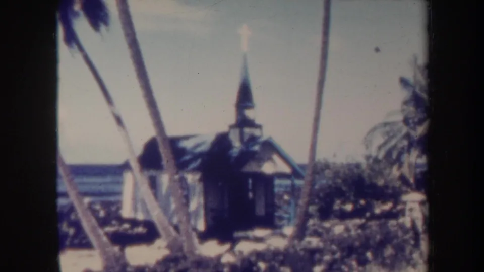 1967 Honolulu-area 8mm frame showing the small wooden church, steeple cross, palm trees, beach vegetation, and ocean backdrop.