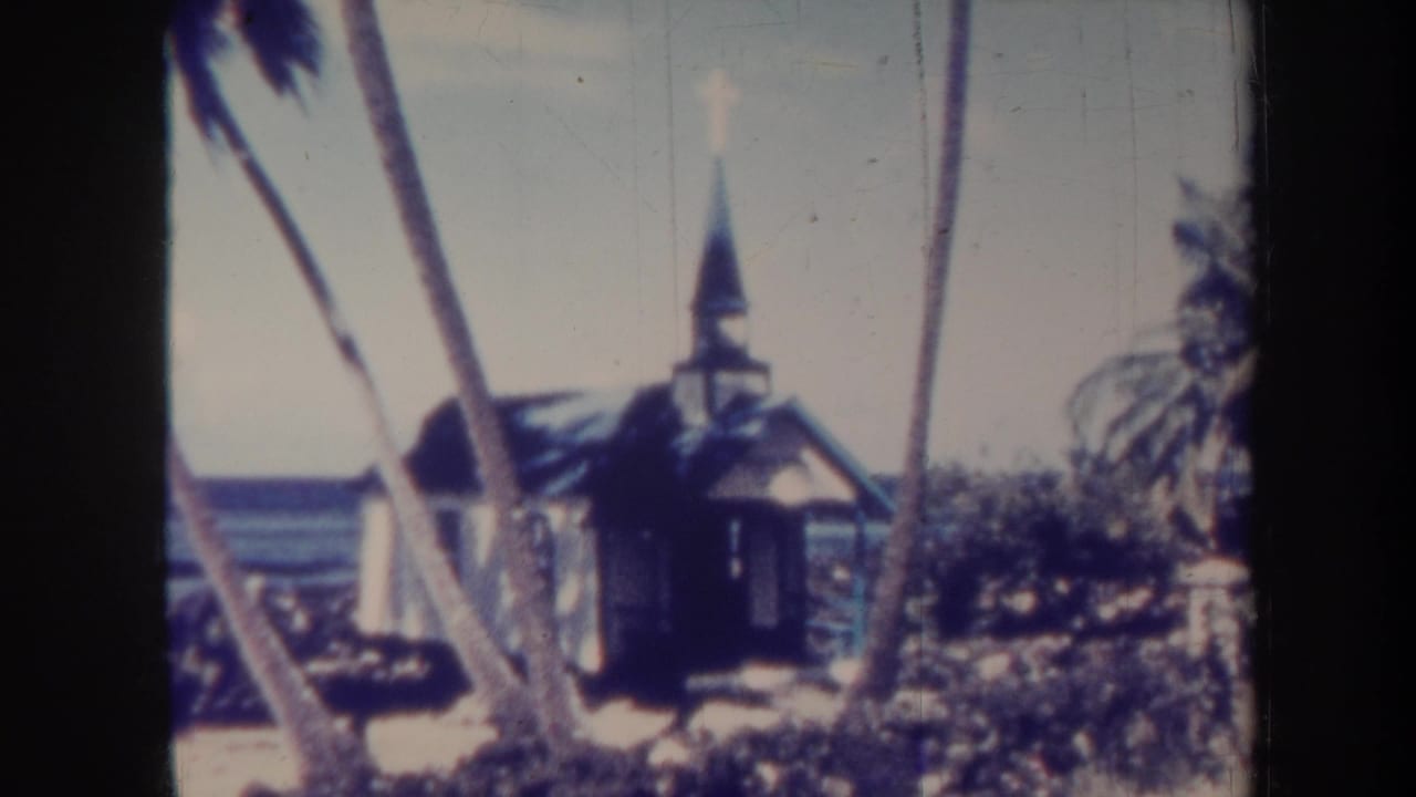 1967 Honolulu home-movie frame showing a small chapel with steeple and cross behind palm trees near the ocean.