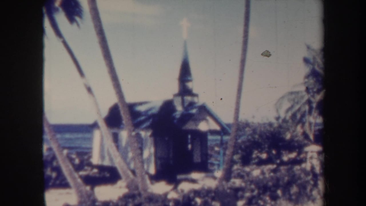 Small chapel with steeple and cross among palms, with blue ocean visible behind.