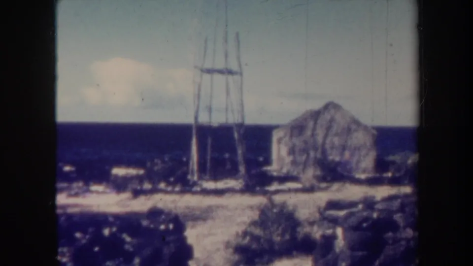 1967 Honolulu coastal view with a rustic low structure, tall wooden frame, ocean horizon, and rocky foreground.