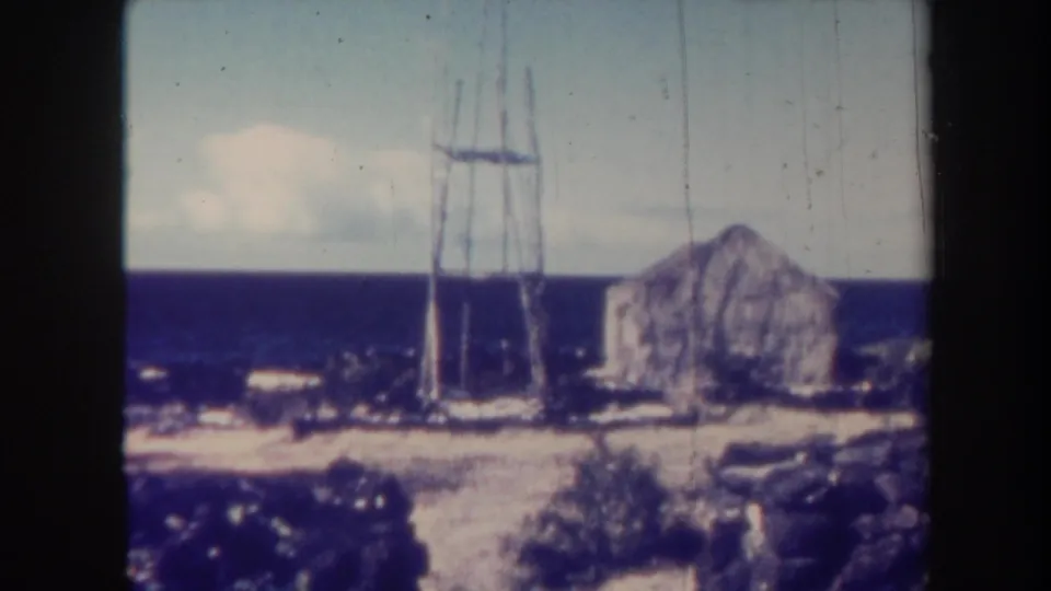Vintage 8mm view of a Honolulu coastal hillside with a tall metal tower, stone building, deep blue sea, and pale cloud cover.