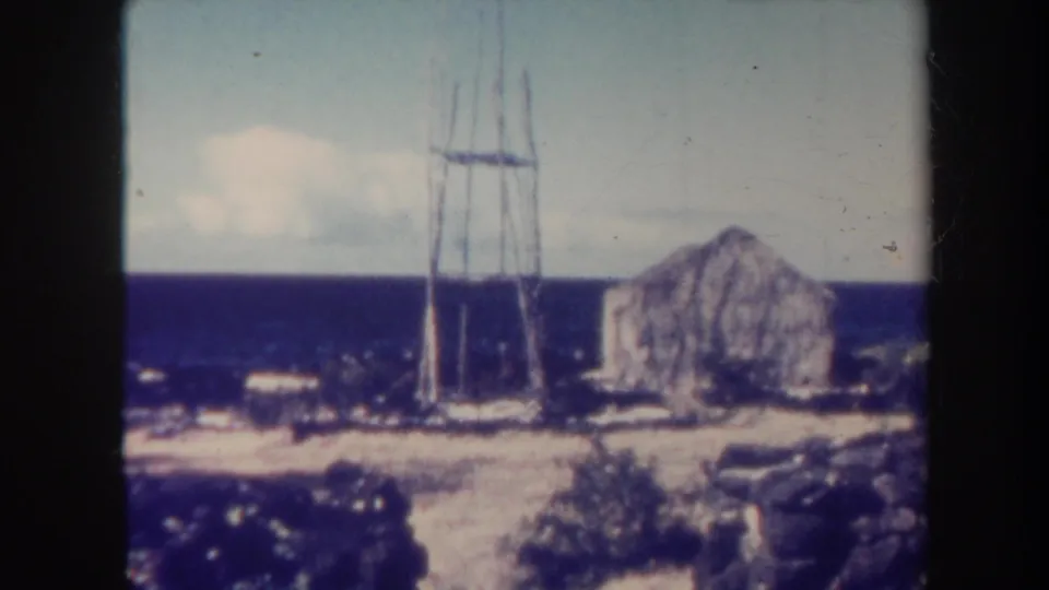 1967 Hawaii home-movie frame showing a stone building, slender utility tower, rough coastal ground, and blue ocean under a bright sky.