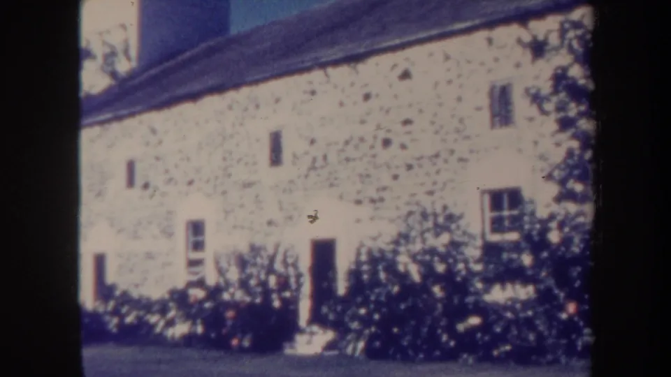1967 Honolulu 8mm frame showing a white masonry exterior, dark roof, doorway, small windows, and planted shrubs along the wall.
