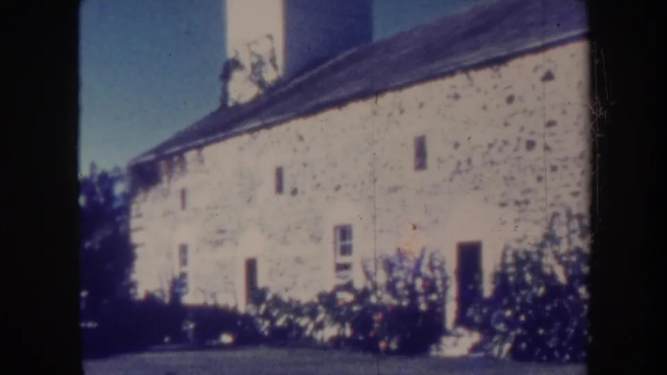 1967 Honolulu 8mm frame with an angled view of the white building, dark roof, small windows, doorway, shrubs, and blue sky.