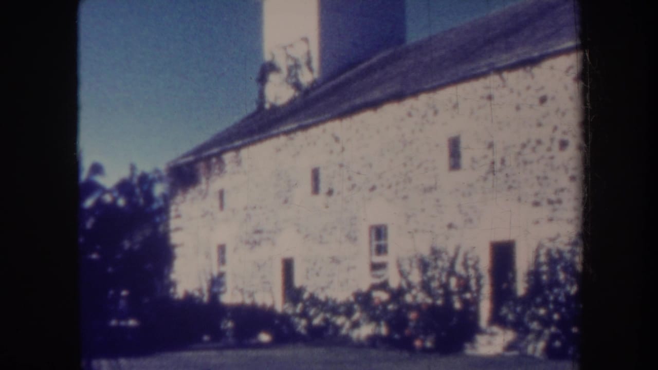 1967 Honolulu 8mm frame showing a pale stone building facade with a dark roofline, small windows, doorway, and garden foliage.