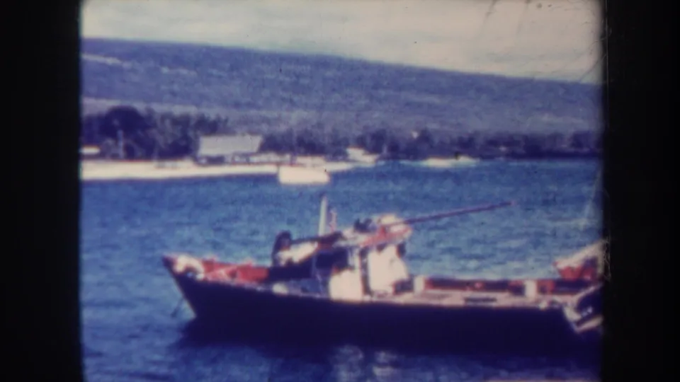 1967 Honolulu home-movie frame showing a small fishing boat on calm coastal water with shoreline buildings and hills in the distance.