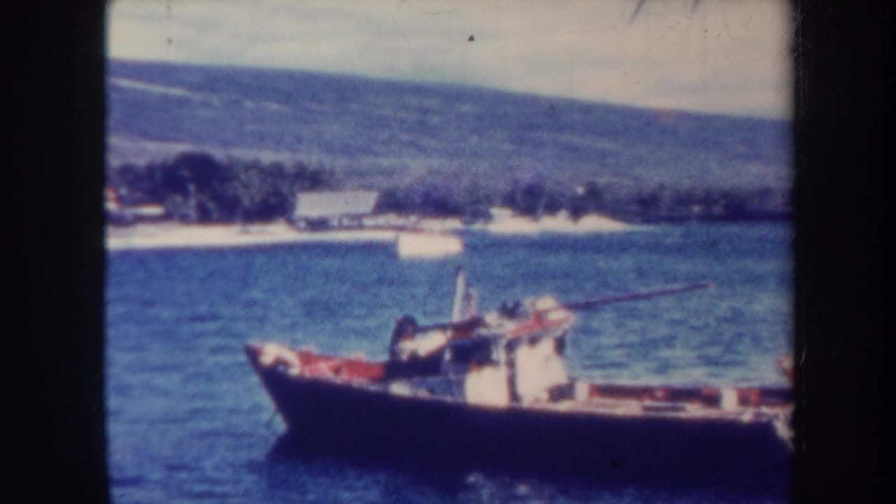 1967 Honolulu home-movie frame showing a small dark boat on calm water with a tropical shoreline and green hills beyond.