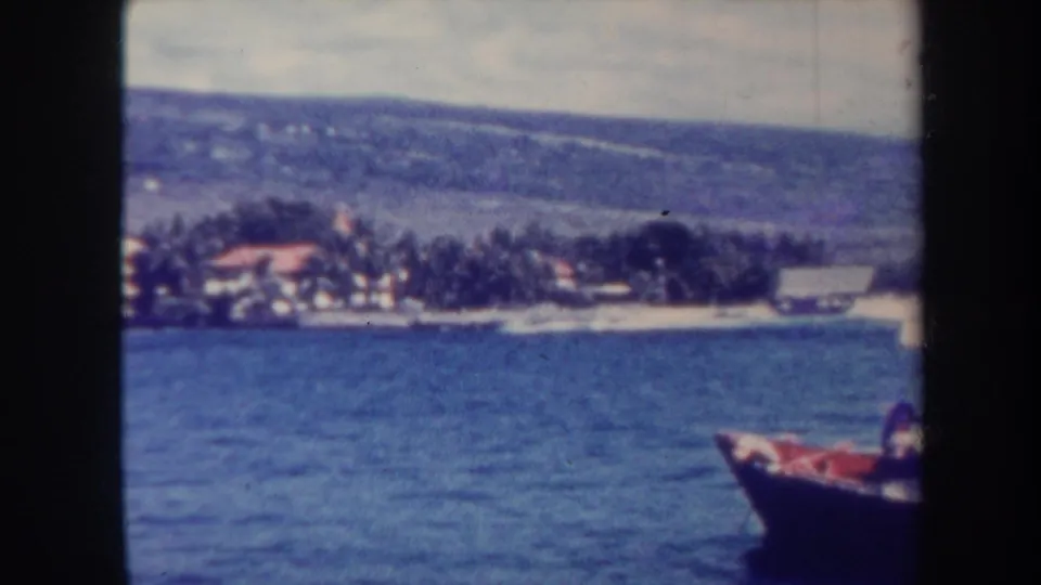 1967 Honolulu home-movie view across calm water toward palms, low buildings, hills, and the edge of a small boat.
