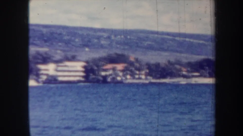 1967 Honolulu frame looking over blue water toward shoreline buildings, palm trees, and green hills under a pale sky.
