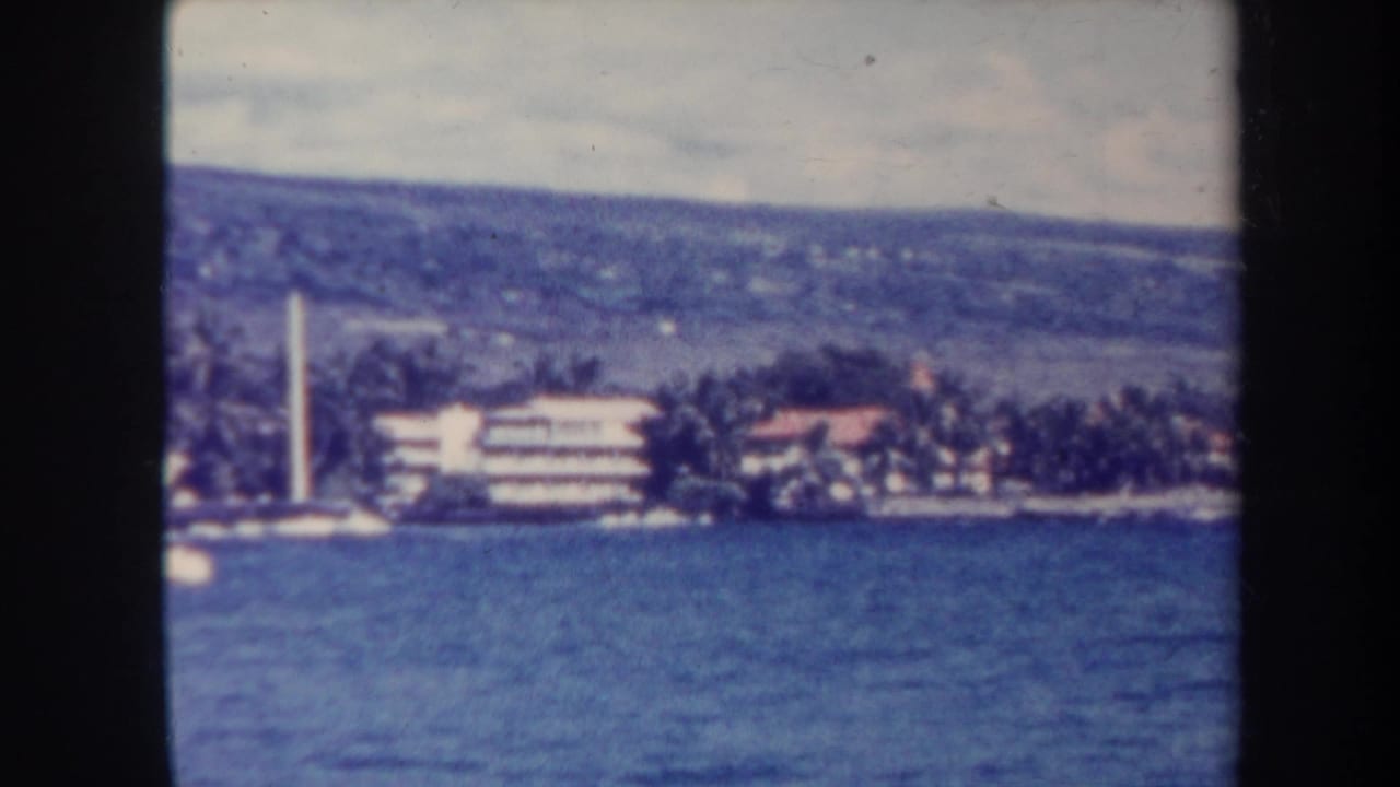 Distant Honolulu shoreline with ocean water, palms, low buildings, hillside, and a white vertical tower.
