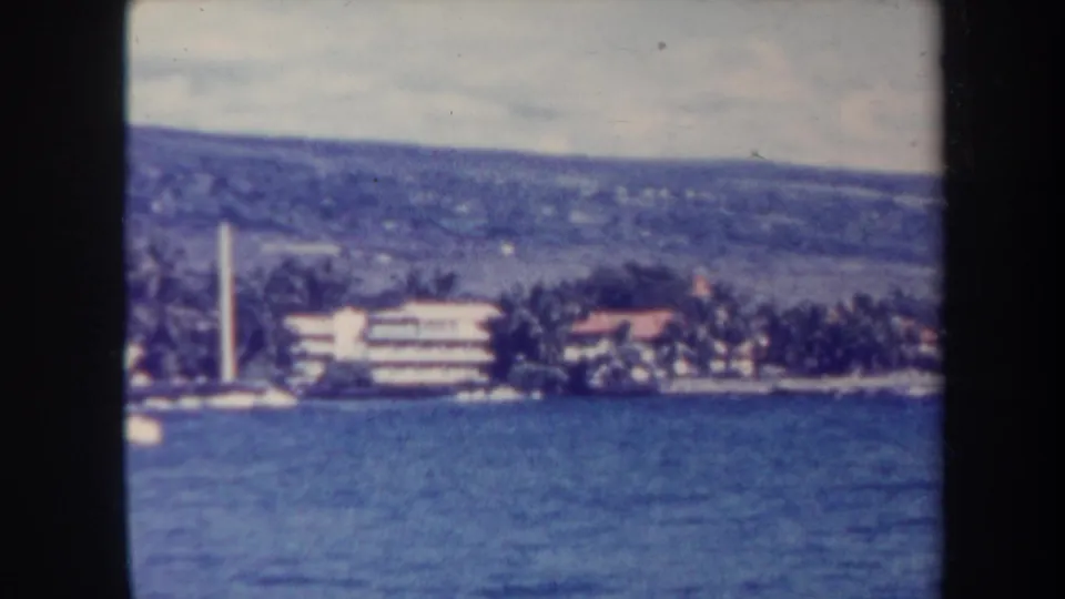 1967 8mm view from the water toward Honolulu shoreline with palms, coastal buildings, hillside, and a white tower-like structure.
