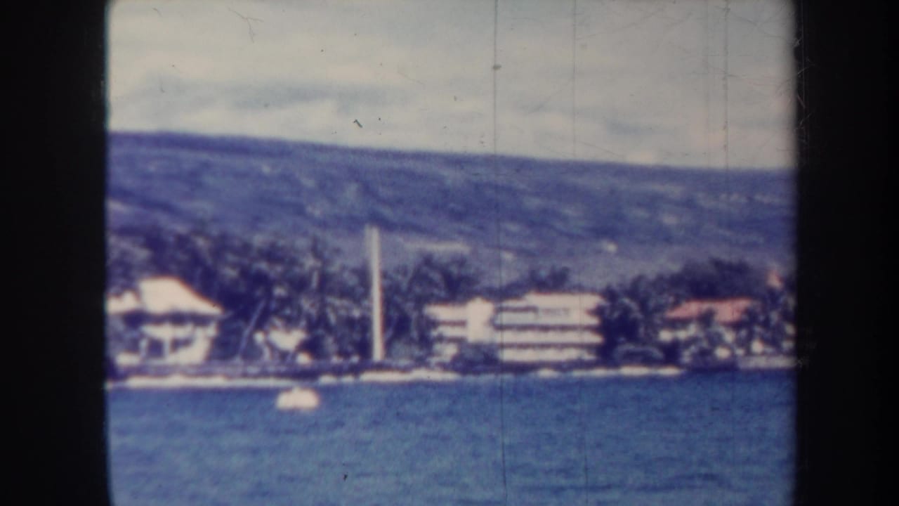 Waterfront buildings and palms along the shore, with a white tower and hillside in the background.