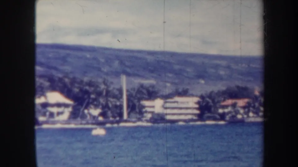 Honolulu waterfront seen from a boat in 1967, showing palms, low buildings, a white vertical tower, water, and hillside.