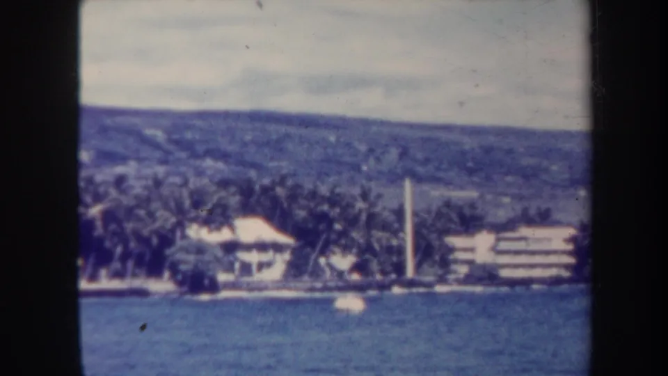 1967 Honolulu shoreline from offshore, with ocean foreground, palms, a light-roofed structure, white tower, and hillside beyond.