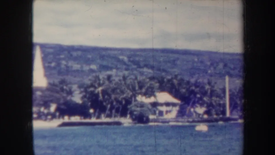 1967 8mm boat-view frame of Honolulu shore with a sailboat, palms, low buildings, a white tower, and hillside.
