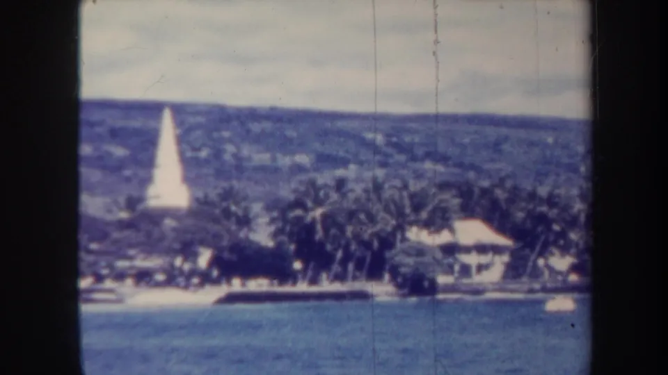 1967 Honolulu home-movie frame showing blue water, a tropical shoreline, palms, small buildings, and a white obelisk on the hillside.