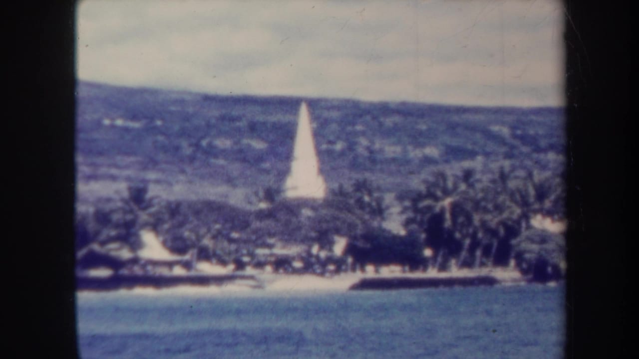 White obelisk above a palm-lined waterfront with calm water and distant hillside.