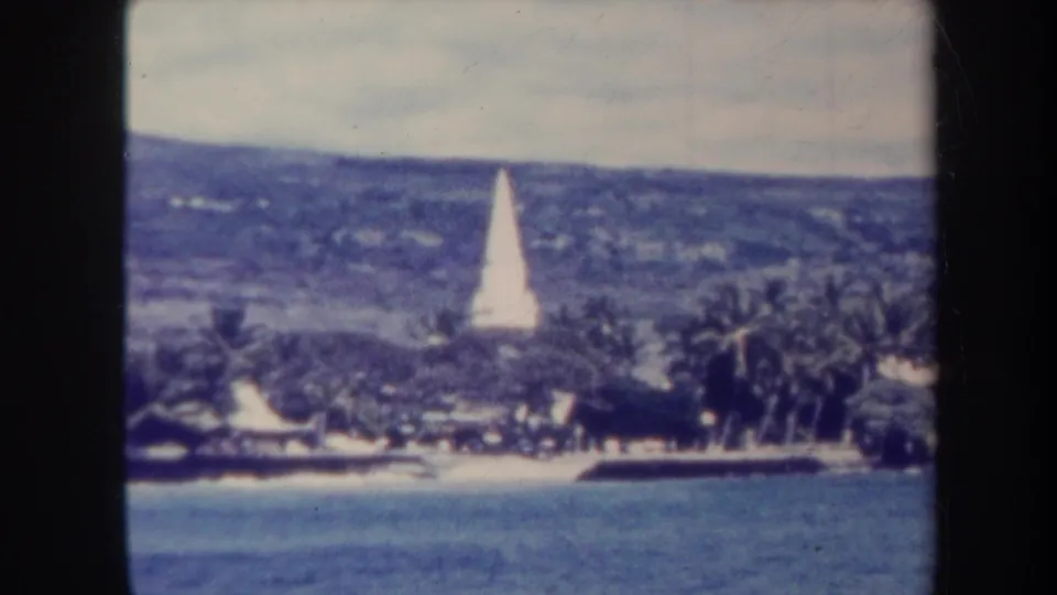 1967 Honolulu coastal view with a white hillside obelisk, palms, low waterfront structures, and soft 8mm film texture.