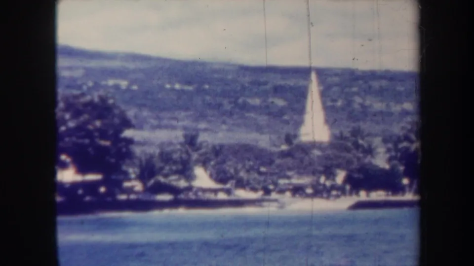 1967 Honolulu 8mm frame showing the shoreline from across the water, with palms, low structures, and a white obelisk near the hillside.