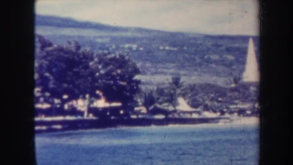1967 Honolulu coastal landscape with calm water, dense shoreline vegetation, small buildings, and a white obelisk at the right of frame.