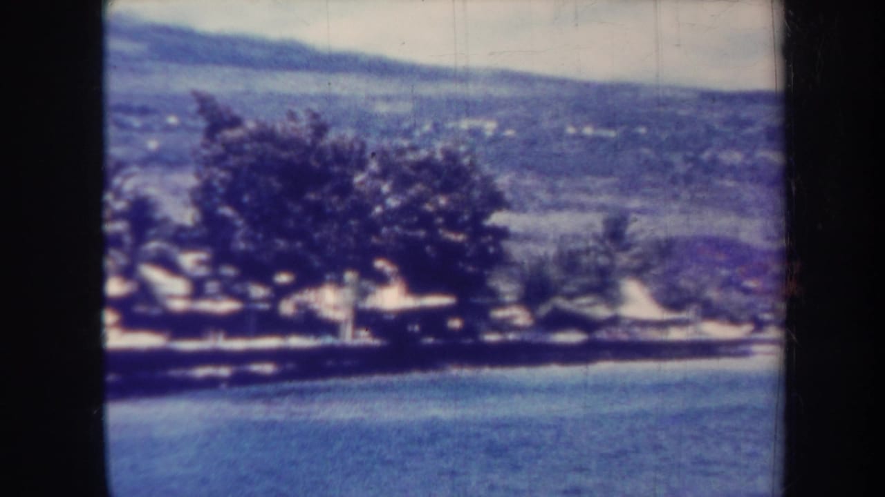 1967 Honolulu 8mm frame showing calm water, shoreline trees, distant hillside, and a soft blue color cast.