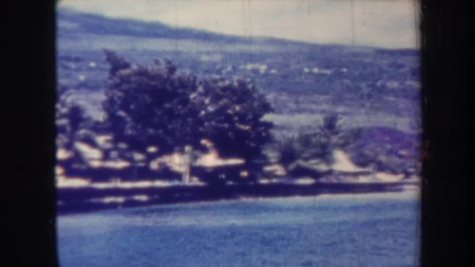 1967 Honolulu 8mm frame showing calm water, shoreline trees, distant hillside, and a soft blue color cast.