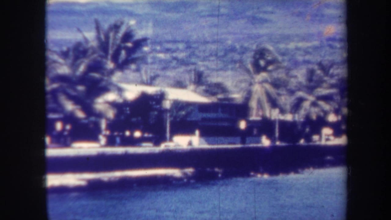 1967 Honolulu home-movie frame showing waves below a seawall, palm trees, low houses, and distant hills with a blue aged-film cast.