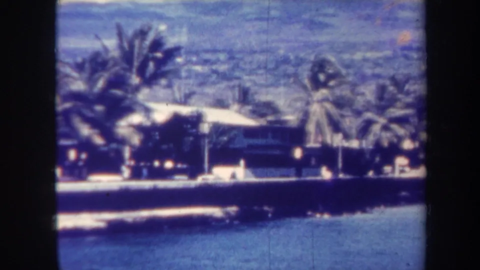 1967 Honolulu home-movie frame showing waves below a seawall, palm trees, low houses, and distant hills with a blue aged-film cast.