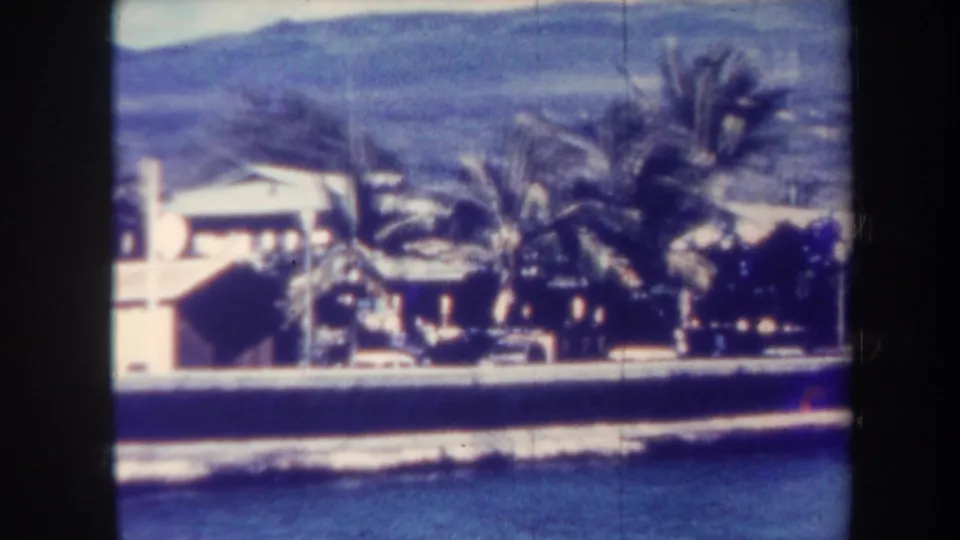 1967 Honolulu home-movie still showing surf below a coastal wall, palms, parked cars, and light houses against a mountain backdrop.