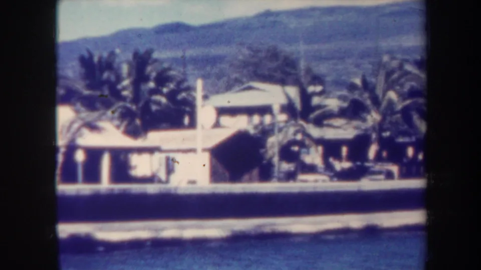 1967 Honolulu frame with a bright shoreline house, palms, a low seawall, breaking surf, and hazy hills beyond.
