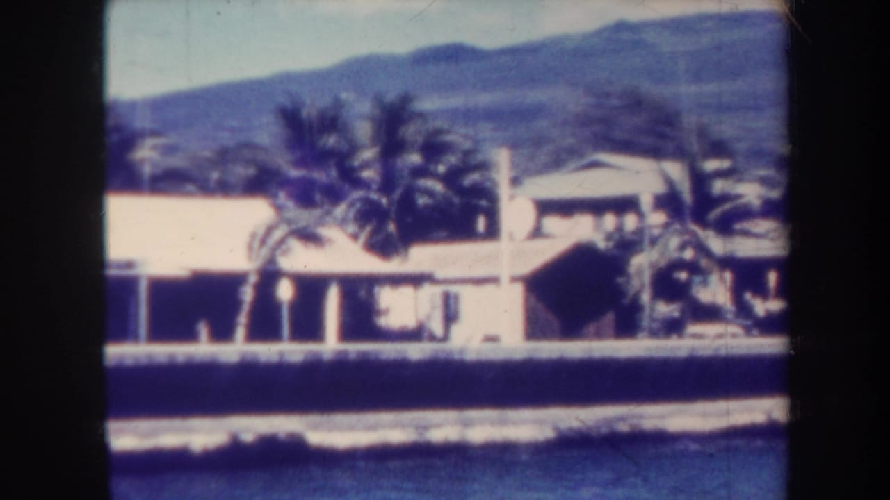 1967 Honolulu 8mm frame showing a low waterside building, palm trees, distant mountains, and a dark band of water in front.