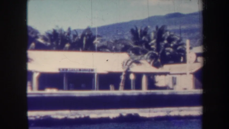 1967 Honolulu frame with a low waterfront building, palms, a partially legible sign, foreground water, and hazy mountains.