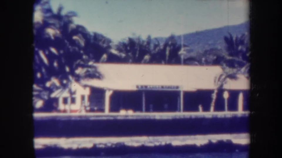 1967 Honolulu 8mm frame showing a broad low building along the water, surrounded by palms with mountains in the distance.
