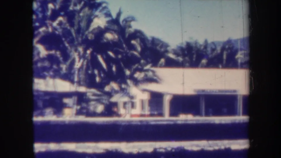 1967 Honolulu frame showing palms in front of a low waterside building, with foreground water and mountains beyond.