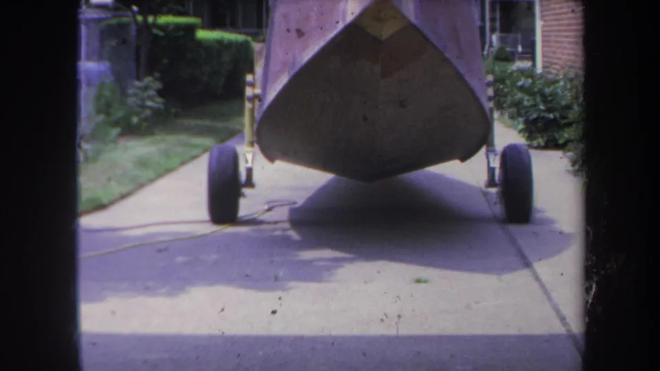 The camera moves nearer to the boat's bow on its trailer, with driveway pavement, hedges, and a residential exterior behind it.