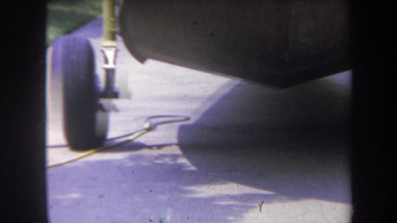 1969 Yucaipa-area 8mm frame showing a dark trailer wheel, metal landing gear, driveway pavement, and a yellow drop cord.