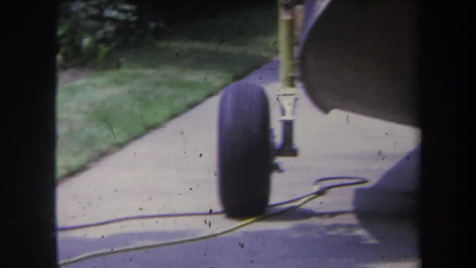 1969 home-movie frame with the trailer wheel closer to center, bordered by driveway, grass, foliage, and visible film grain.