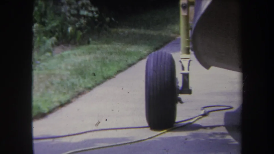 Yucaipa-area 8mm frame showing a trailer wheel and landing gear over a driveway, with a yellow cord, lawn, and shrubs behind it.