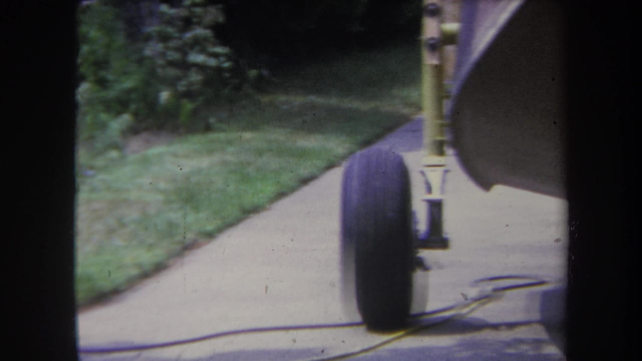 1969 Yucaipa home-movie frame showing a small aircraft wheel and landing gear beside a driveway and lawn.