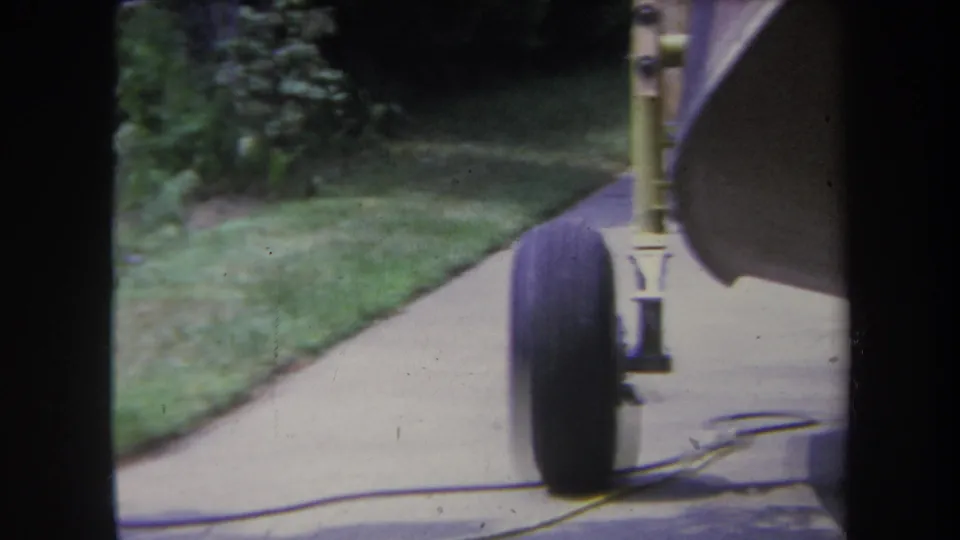 1969 Yucaipa home-movie frame showing a small aircraft wheel and landing gear beside a driveway and lawn.