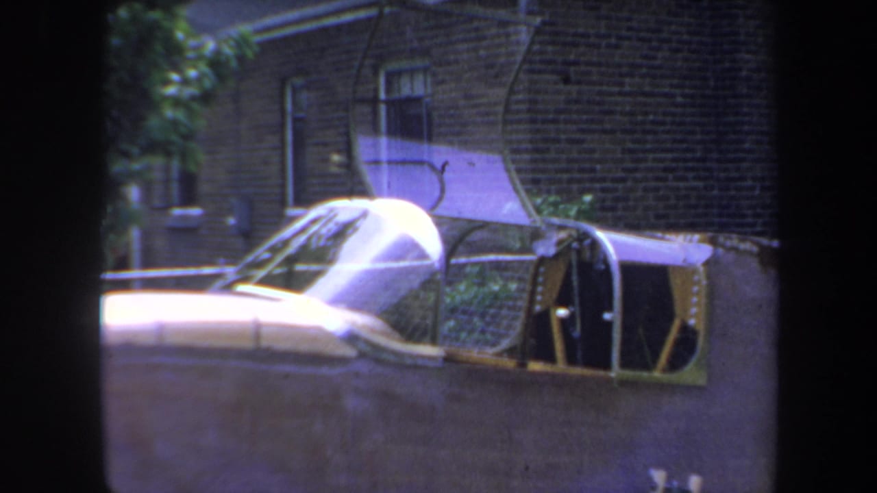 Small aircraft with bubble canopy and open cockpit beside a brick house.