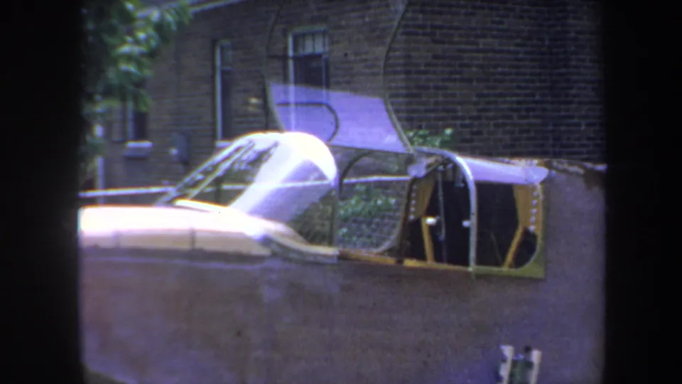 1969 Yucaipa frame with the small aircraft's canopy, open cockpit structure, wing, and brick house background.