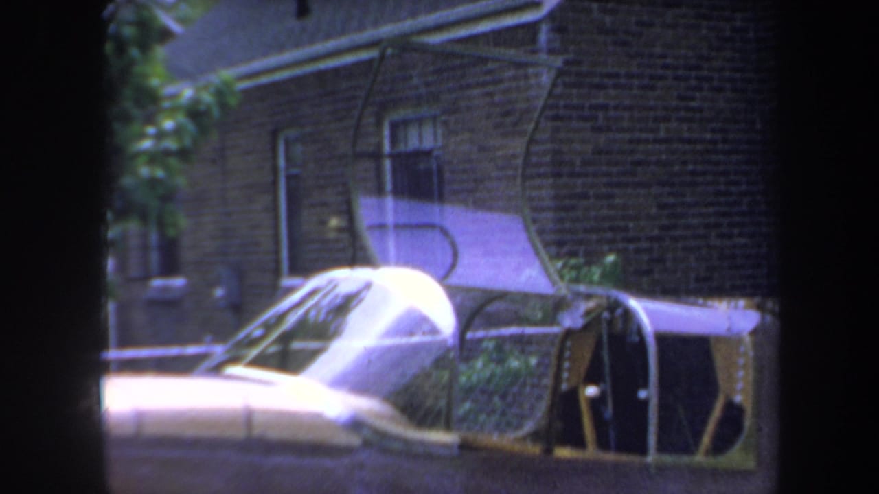 1969 Yucaipa-area 8mm frame showing a gold-colored classic car beside a brick building with windows and reflections.