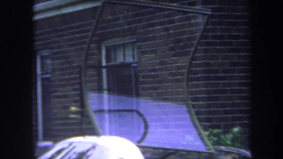 1969 Yucaipa-area 8mm frame with a brick wall, windows, curved foreground lines, and the roofline of a gold-colored car.
