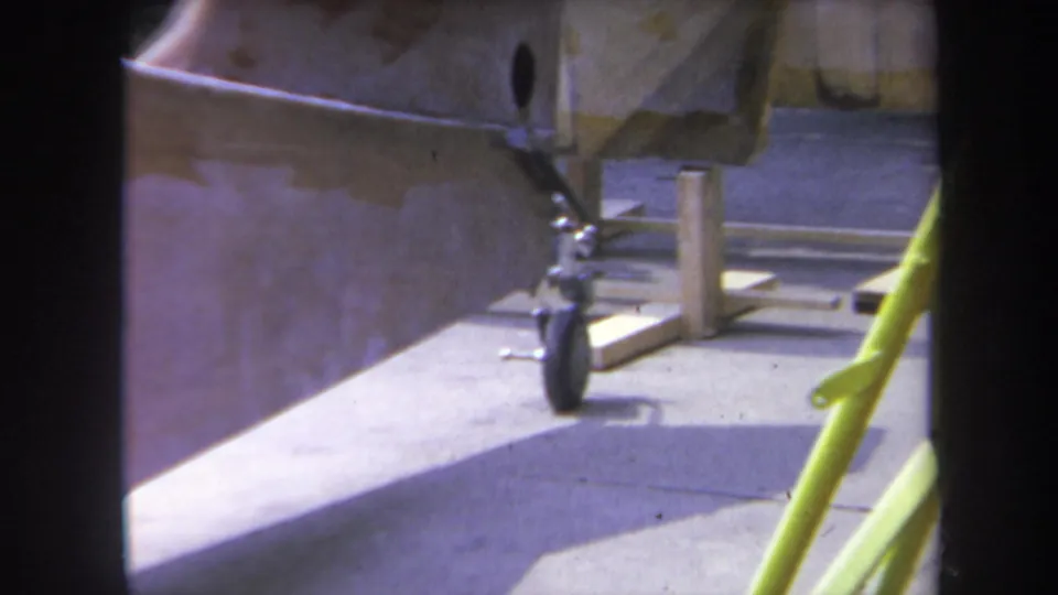 1969 Yucaipa frame showing the underside of a parked small aircraft with landing wheel, wooden supports, pavement, and partial strut.