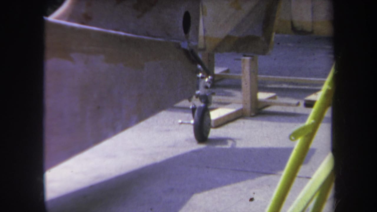 1969 Yucaipa home-movie frame showing an unpainted aircraft rear section, landing gear, wood supports, and a yellow foreground pole.