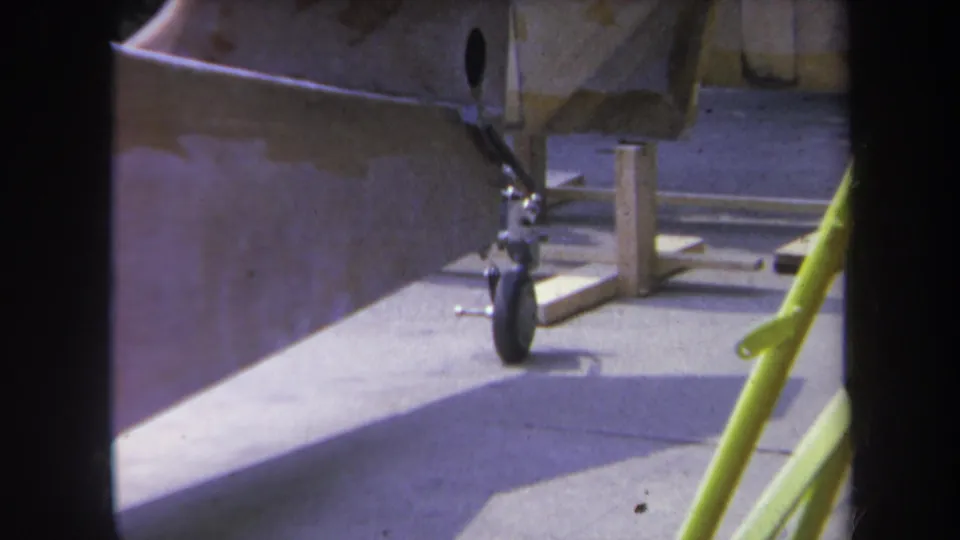 1969 Yucaipa home-movie frame showing an unpainted aircraft rear section, landing gear, wood supports, and a yellow foreground pole.