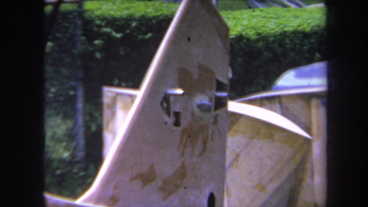 Metal machine part with stained surface, projecting joint, fence, and hedge in the background.