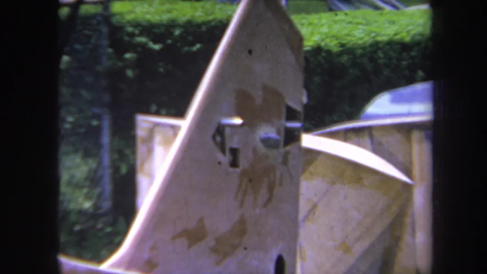 Close 1969 Yucaipa frame of a stained industrial metal part with a protruding joint, chain-link fence, and green hedge behind it.