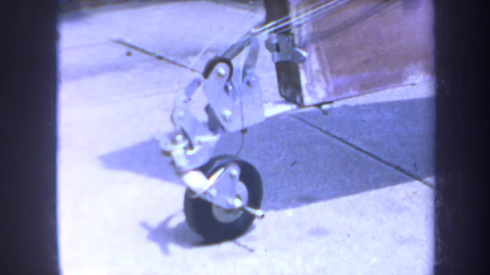 Final 1969 8mm frame showing a close-up of a model airplane wheel, metal landing gear, and struts on a lighter surface.